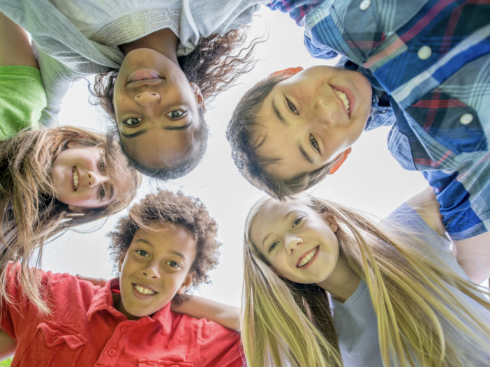 Smiling group of school-age children looking down at the camera in a circle, symbolizing family and back-to-school season.
