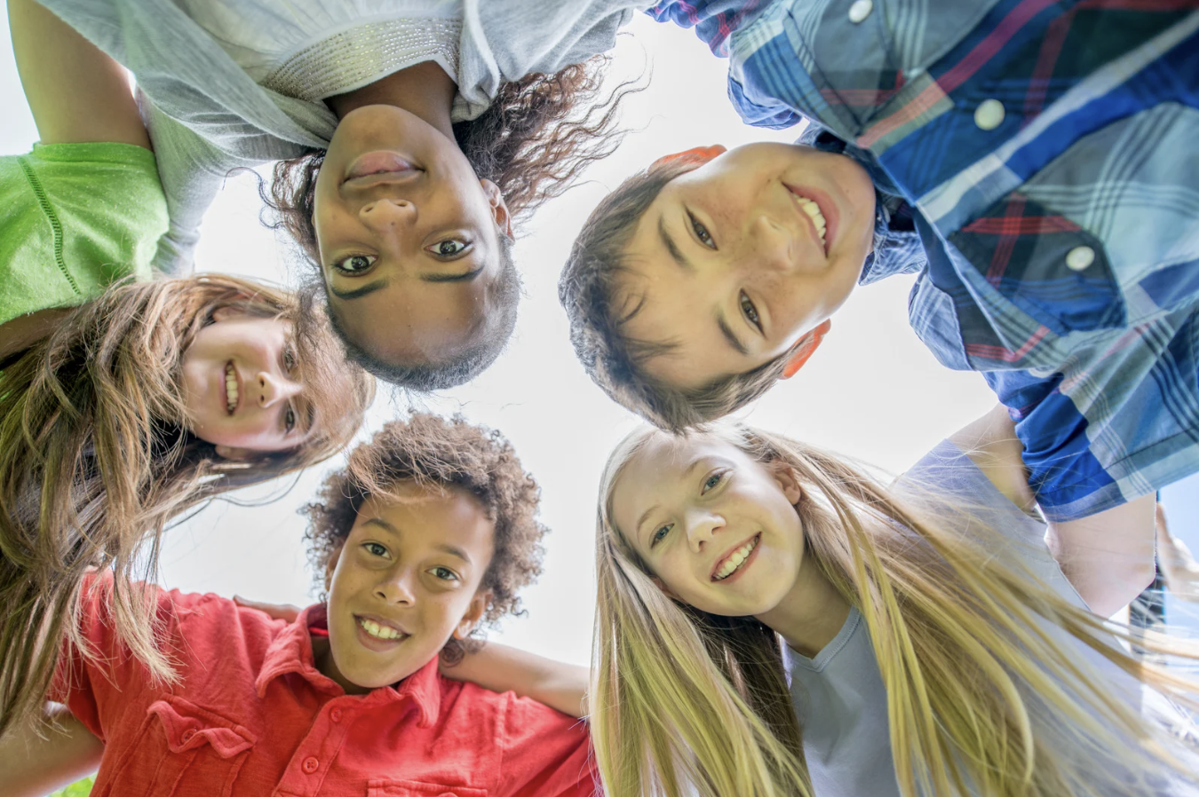 Smiling group of school-age children looking down at the camera in a circle, symbolizing family and back-to-school season.