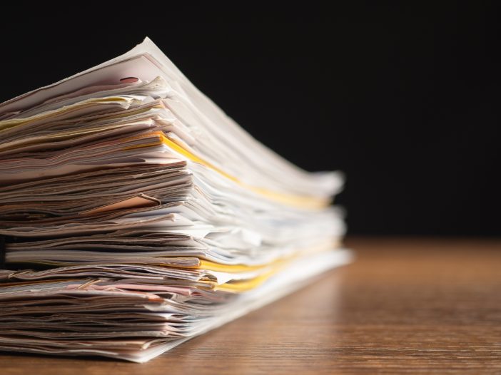 Close-up of a tall stack of old papers and folders sitting on a wooden table against a dark background.