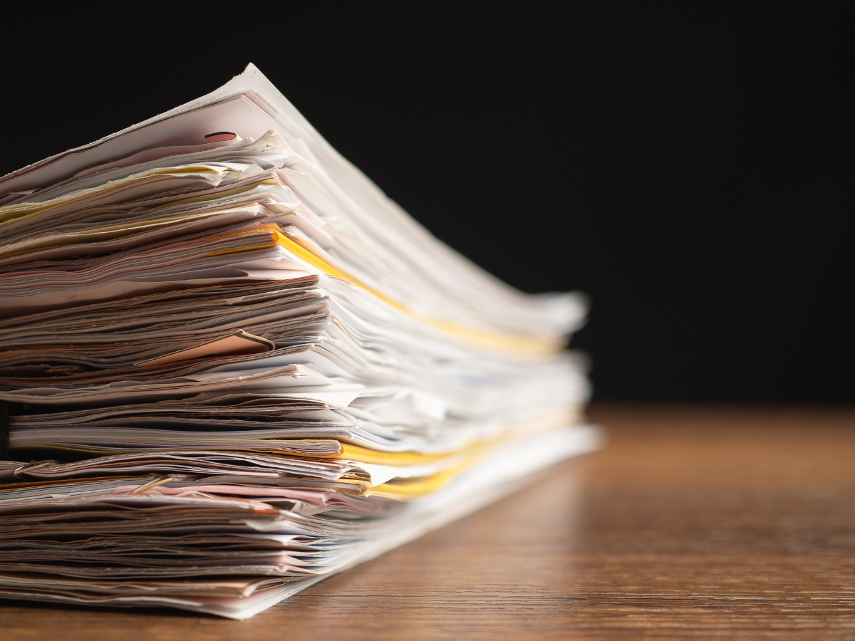 Close-up of a tall stack of old papers and folders sitting on a wooden table against a dark background.
