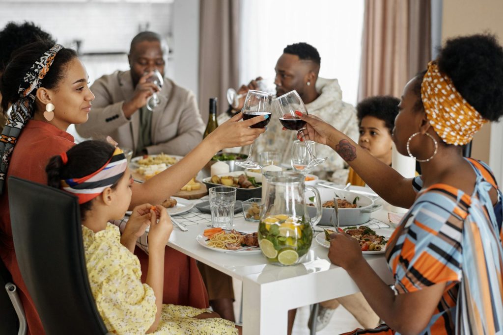 A family shares a meal together at a bright dining table, with adults raising glasses in a toast while children sit nearby and plates of food fill the table.