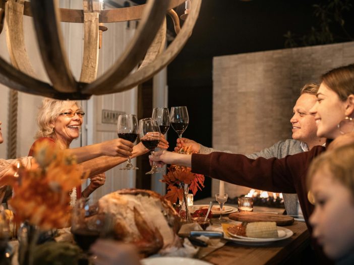 A group of relatives gathered around a warmly lit dinner table clinks wine glasses in a celebratory toast, with a holiday meal and decorations in front of them.