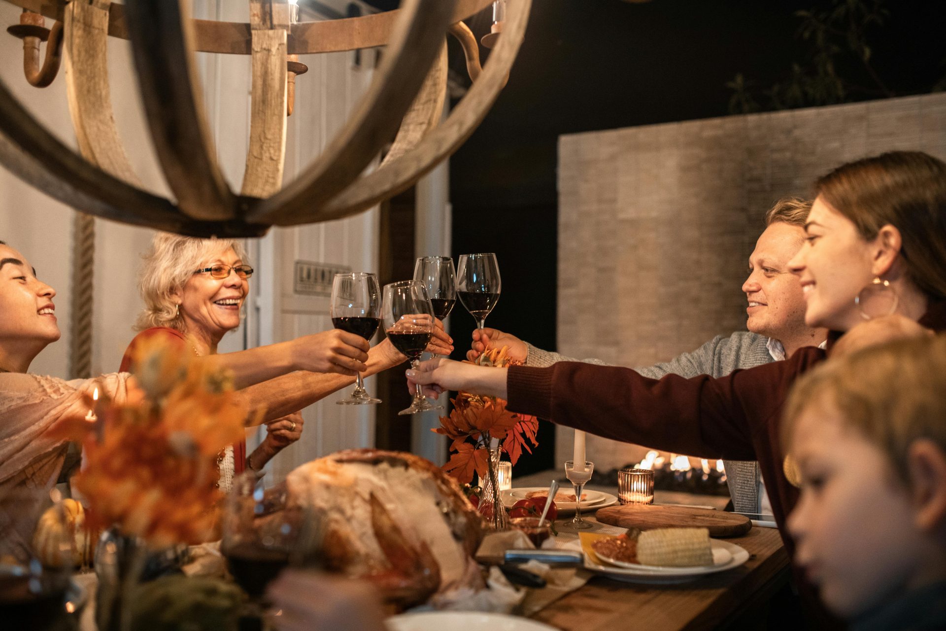 A group of relatives gathered around a warmly lit dinner table clinks wine glasses in a celebratory toast, with a holiday meal and decorations in front of them.