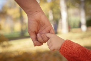 A parent’s hand gently holding a young child’s hand while walking outdoors on a sunny fall day.