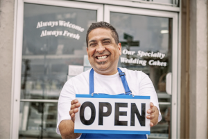 Smiling small business owner wearing an apron and holding an ‘Open’ sign in front of a storefront.