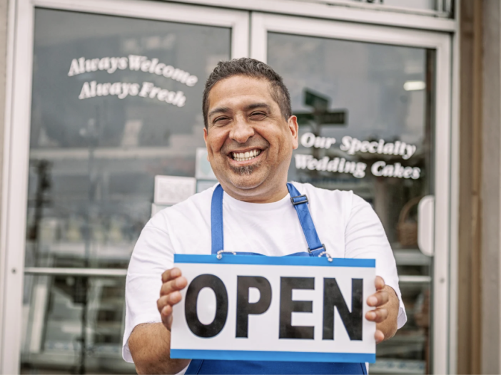 Smiling small business owner wearing an apron and holding an ‘Open’ sign in front of a storefront.