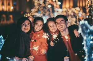 Family of four smiling and holding sparklers during a festive holiday evening with warm lights in the background