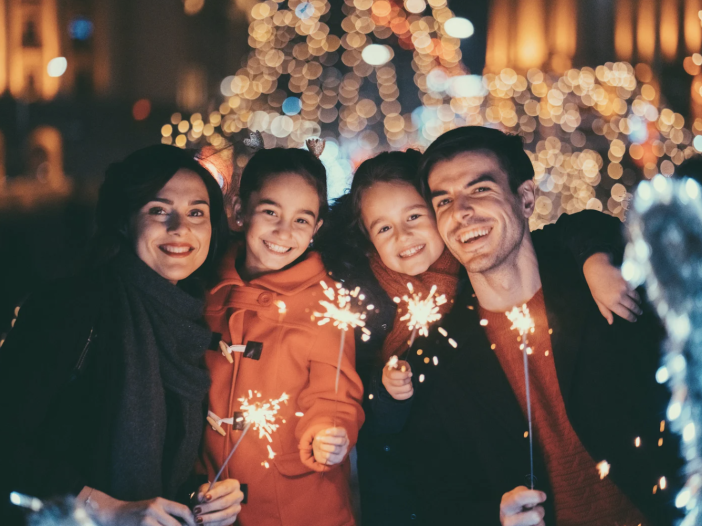 Family of four smiling and holding sparklers during a festive holiday evening with warm lights in the background