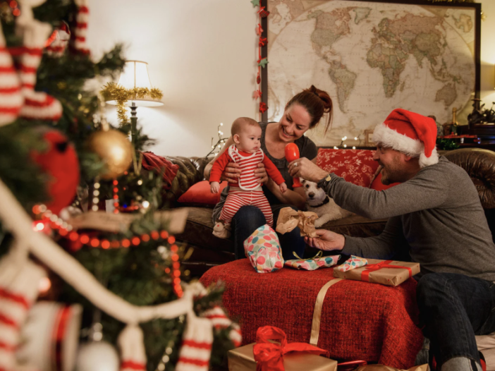 A family with a baby sits together in a cozy living room decorated for Christmas, opening presents near a lit tree and a world map on the wall.