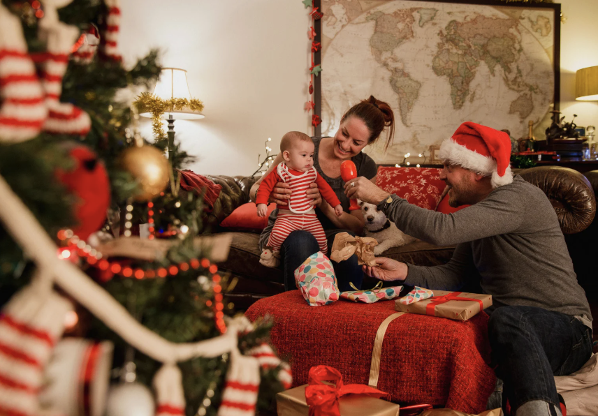 A family with a baby sits together in a cozy living room decorated for Christmas, opening presents near a lit tree and a world map on the wall.