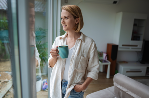 A woman standing by a window at home, holding a coffee mug and looking outside during a quiet moment of reflection