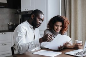 Two adults sitting at a kitchen table reviewing printed documents together with a laptop nearby