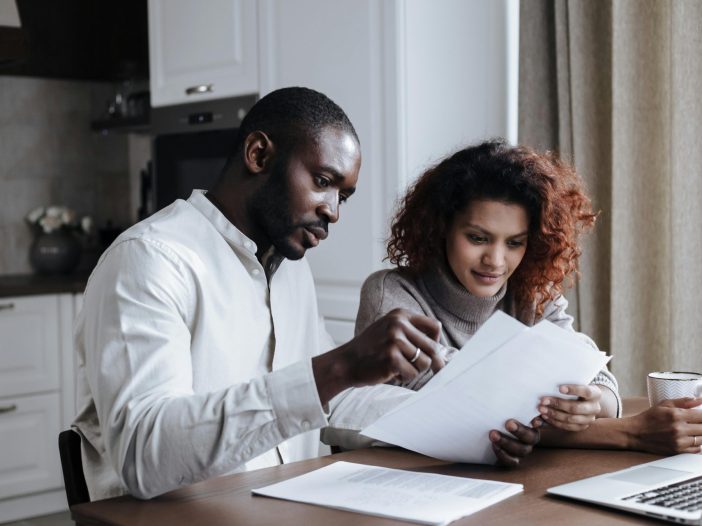 Two adults sitting at a kitchen table reviewing printed documents together with a laptop nearby