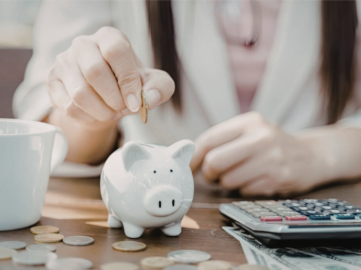 Hand placing coin into white piggy bank next to calculator and coins