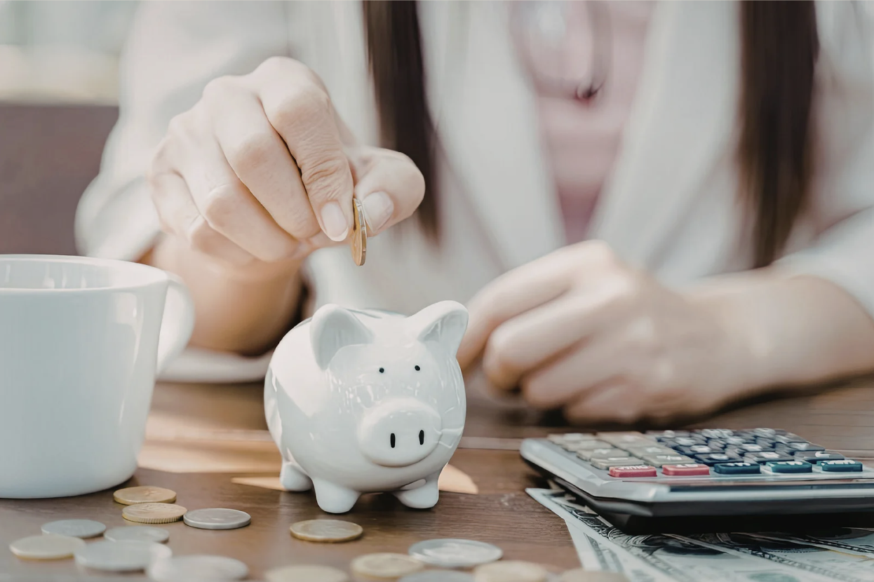 Hand placing coin into white piggy bank next to calculator and coins