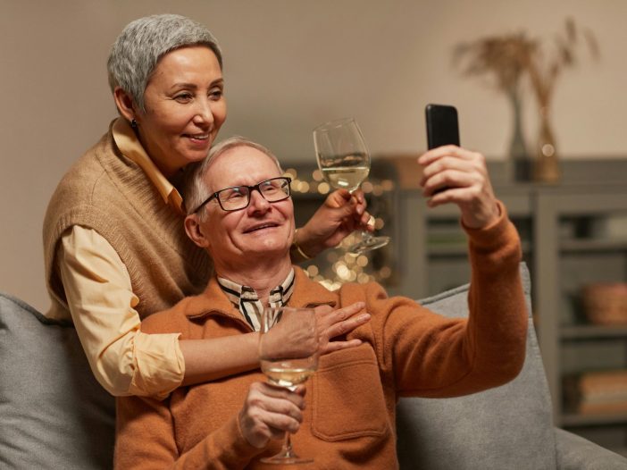 Older unmarried couple sitting together on a couch taking a selfie and holding wine glasses
