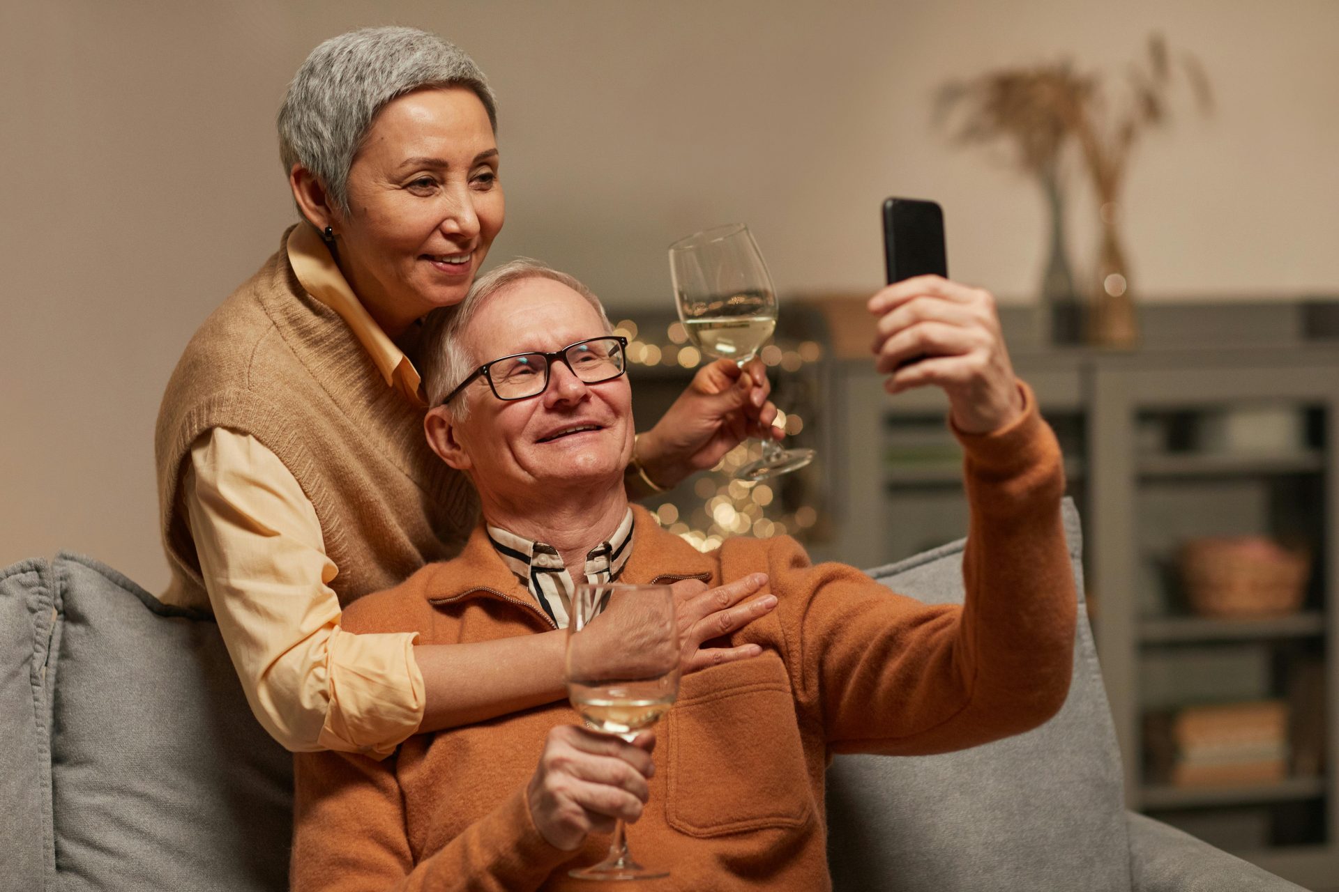 Older unmarried couple sitting together on a couch taking a selfie and holding wine glasses