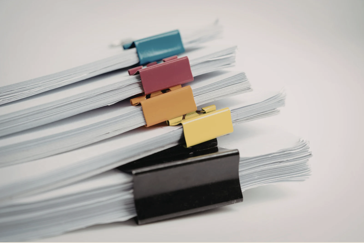 Stacks of white paper documents secured with colorful binder clips on a light gray surface