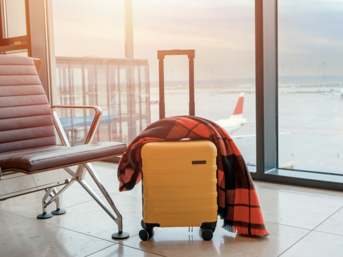 Yellow rolling suitcase with a red plaid blanket draped over the handle, sitting in an airport terminal beside a leather bench with airplanes visible through the window