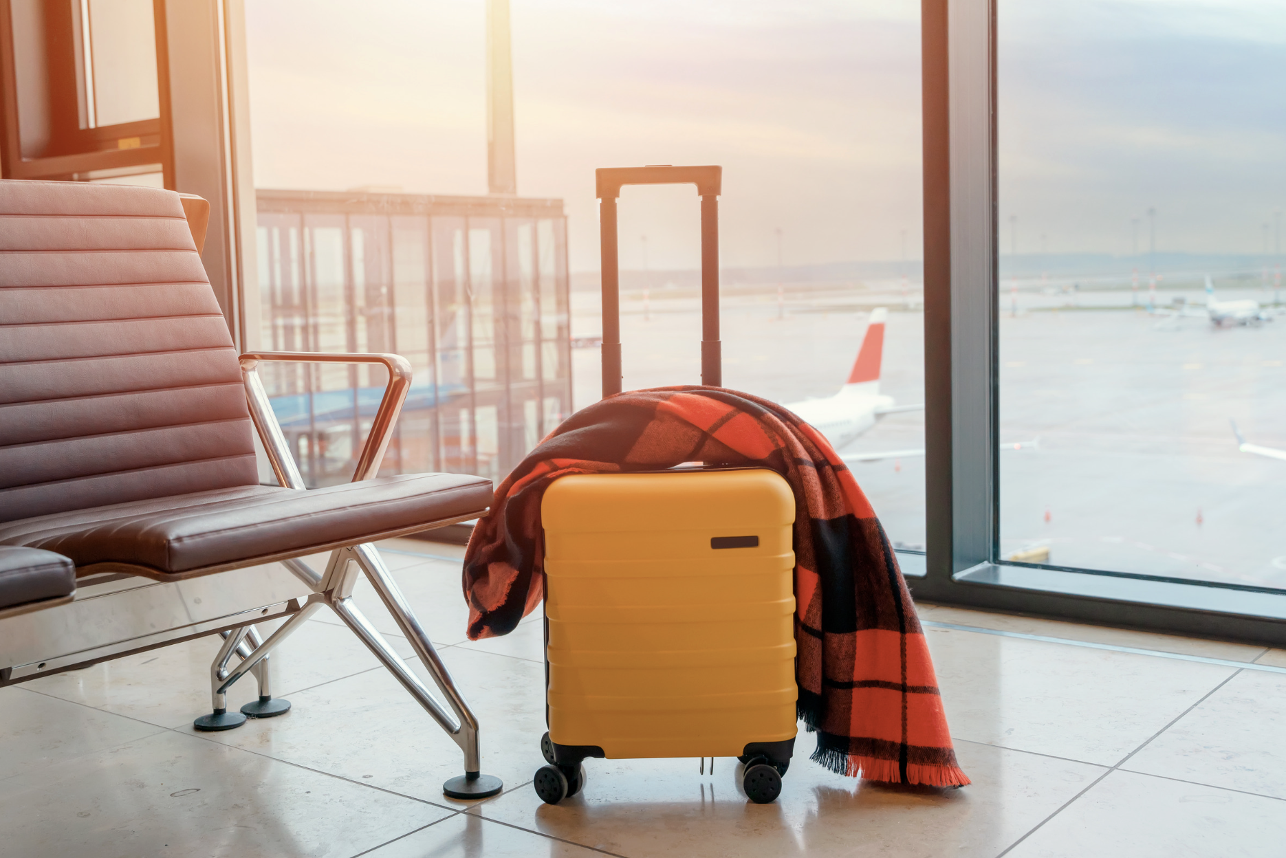 Yellow rolling suitcase with a red plaid blanket draped over the handle, sitting in an airport terminal beside a leather bench with airplanes visible through the window