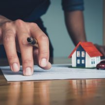 Hand holding a pen near estate planning documents on a desk, with a miniature house and car model resting on the papers