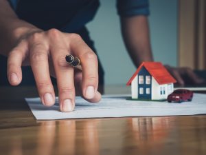 Hand holding a pen near estate planning documents on a desk, with a miniature house and car model resting on the papers