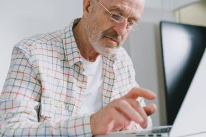 An older gentleman looking at a laptop screen and typing on the keyboard.