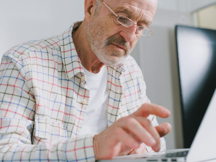An older gentleman looking at a laptop screen and typing on the keyboard.