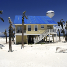 Beach house on stilts with a blue tarp roof surrounded by palm trees on a white sand beach, with a water tower in the background. A vacation home like this one may need a revocable trust or LLC to avoid ancillary probate in more than one state.