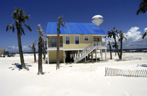 Beach house on stilts with a blue tarp roof surrounded by palm trees on a white sand beach, with a water tower in the background. A vacation home like this one may need a revocable trust or LLC to avoid ancillary probate in more than one state.