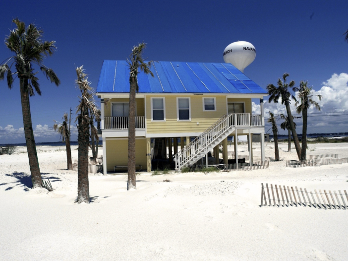 Beach house on stilts with a blue tarp roof surrounded by palm trees on a white sand beach, with a water tower in the background. A vacation home like this one may need a revocable trust or LLC to avoid ancillary probate in more than one state.