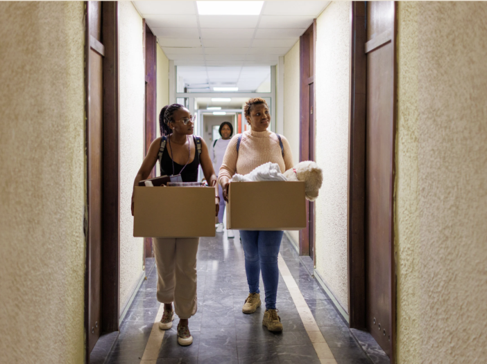 Mother and her 19 year old daughter carrying moving boxes down a college dorm hallway on move-in day