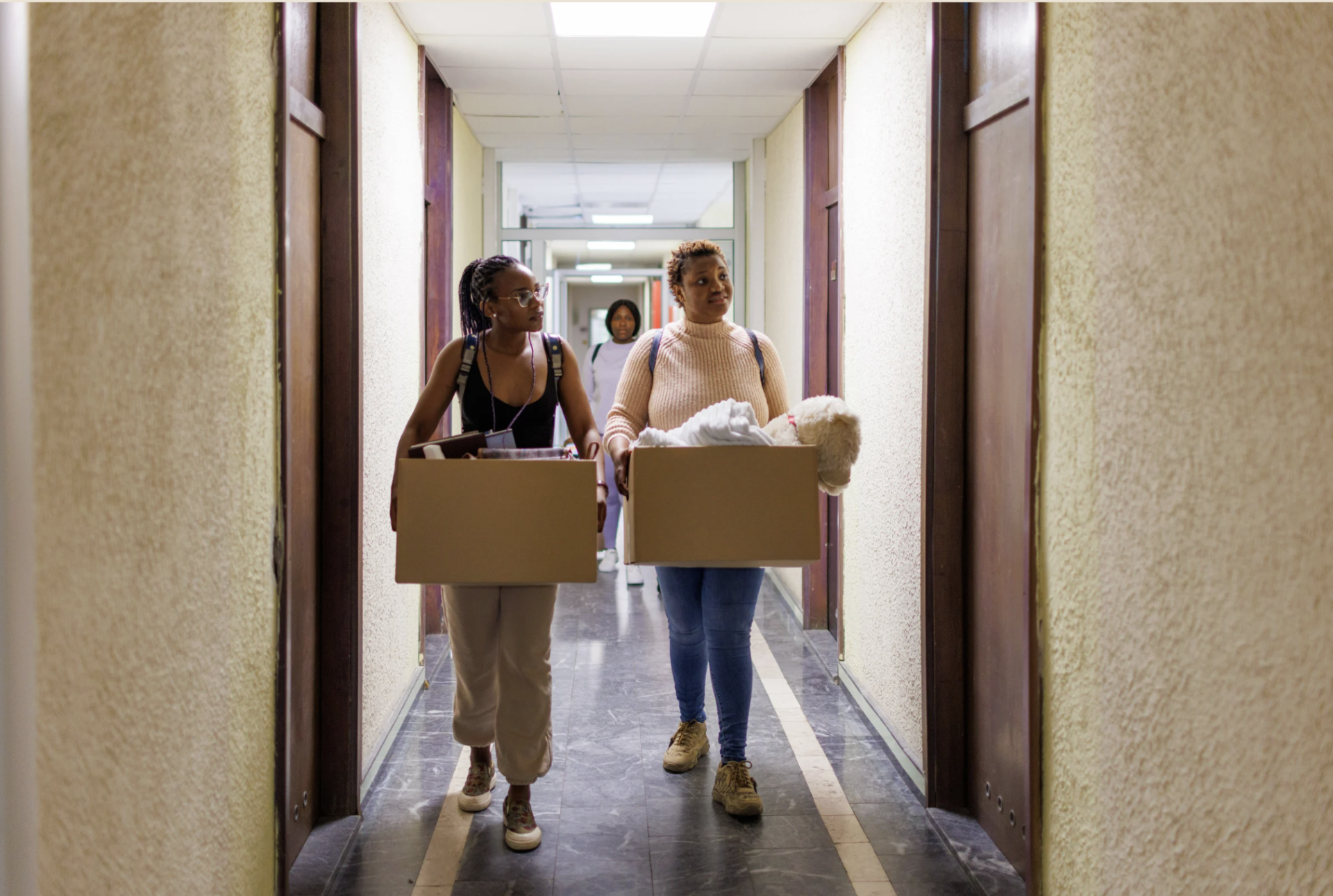 Mother and her 19 year old daughter carrying moving boxes down a college dorm hallway on move-in day