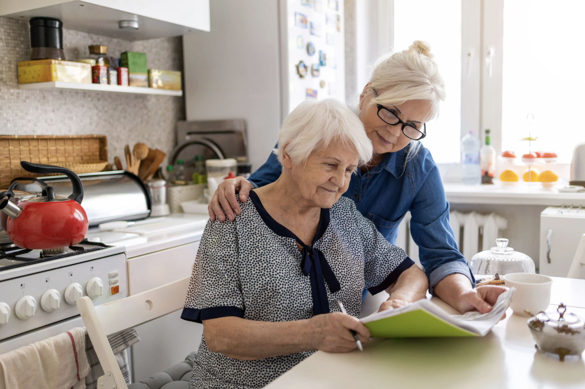 Older mother and adult daughter sitting together reviewing documents at a kitchen table